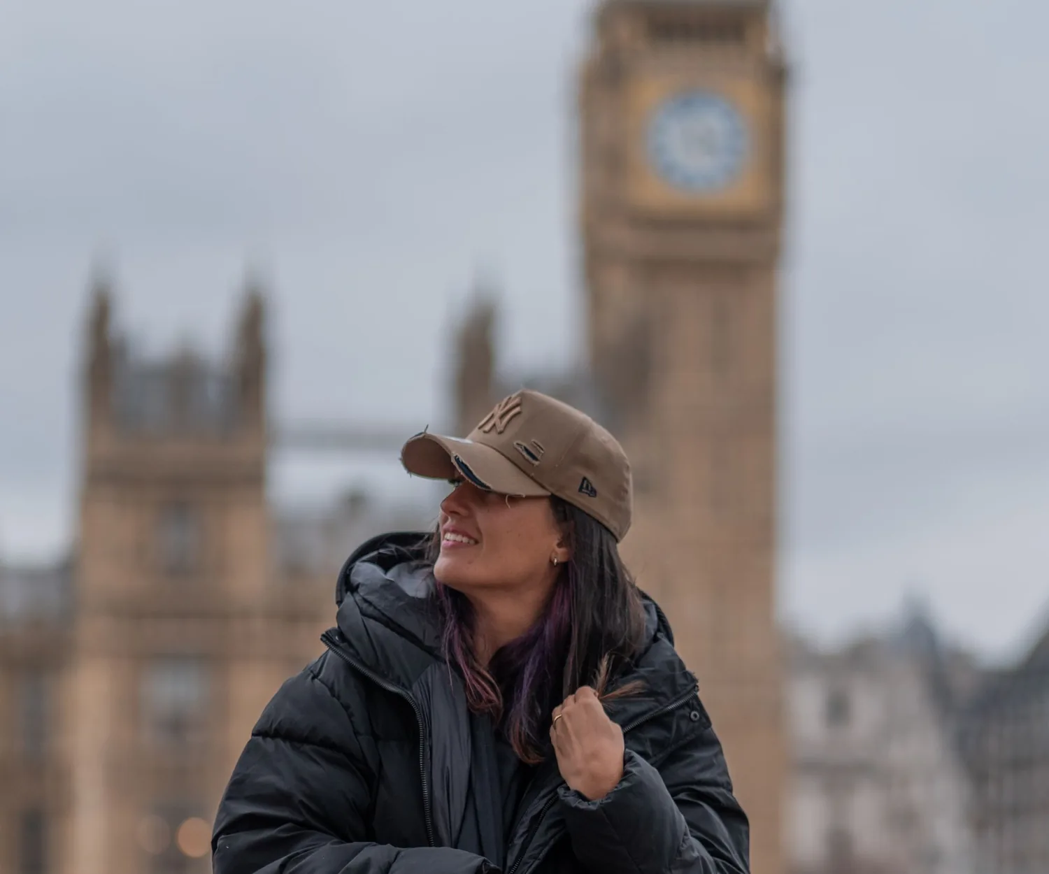 A woman in a dark coat sitting by the Thames with Big Ben and the Houses of Parliament behind her